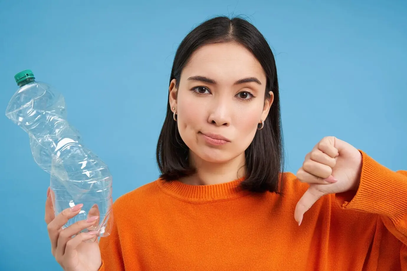 Porträt einer asiatischen Frau, die den Daumen nach unten und eine Plastikflasche zeigt und damit fehlendes Recycling missbilligt, vor blauem Hintergrund.